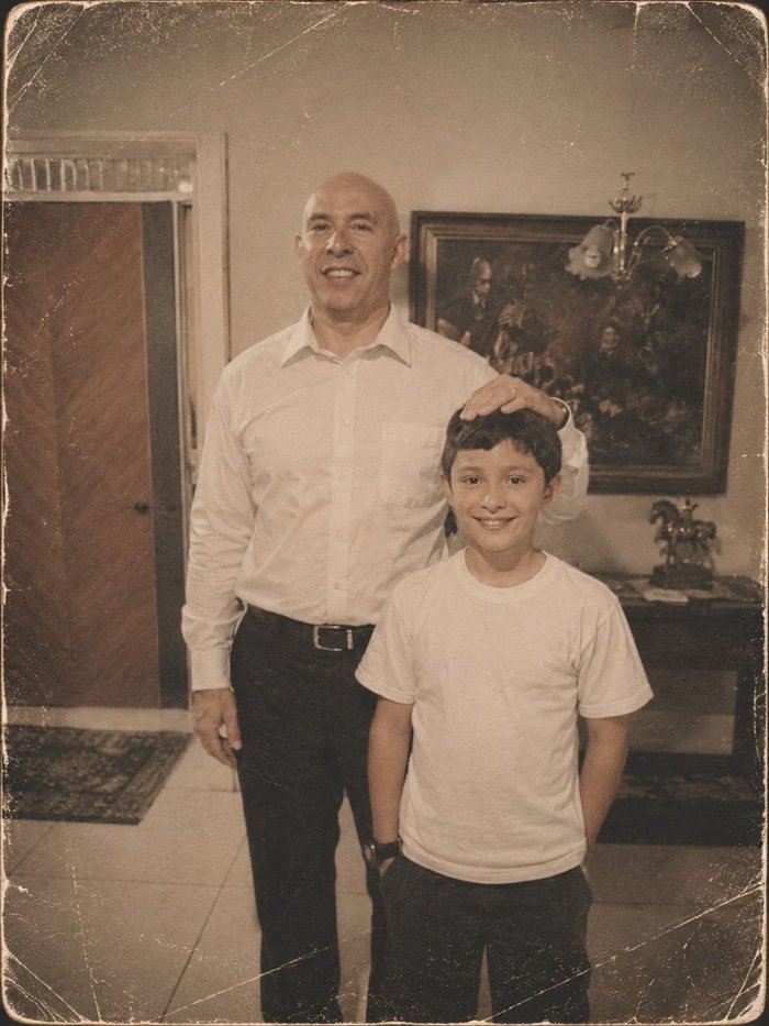 El suegro, calvo y de camisa blanca, con la mano sobre la cabeza de su hijo pequeño, los dos sonriendo en la entrada de la casa